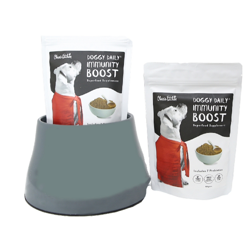 Packaging of 'Olives Kitchen Doggy Daily' showing a white bag with red accents and a picture of a dog bowl. A gray bowl with a supplement inside is also displayed in front of the bag.